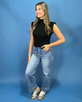 A woman modeling a black sleeveless mock neck top and light-wash blue jeans against a solid blue studio backdrop.