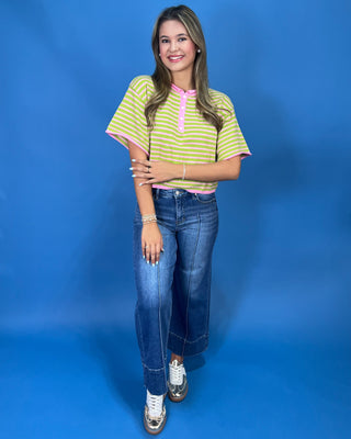 A woman modeling a short-sleeve striped henley top with lime green and white horizontal stripes and pink trim, paired with blue jeans, against a solid blue studio background.