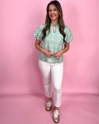 Woman wearing a green patterned top and white pants on a pink background