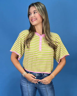 A woman modeling a short-sleeve striped henley top with lime green and white horizontal stripes and pink trim, paired with blue jeans, against a solid blue studio background.