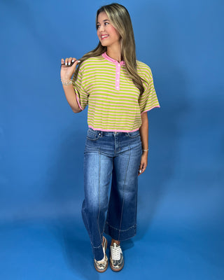 A woman modeling a short-sleeve striped henley top with lime green and white horizontal stripes and pink trim, paired with blue jeans, against a solid blue studio background.