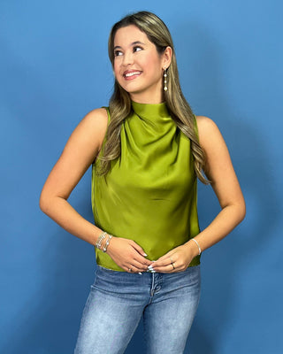 A woman wearing a sleeveless high-neck satin top in olive green, paired with blue jeans, posing against a solid blue studio background.
