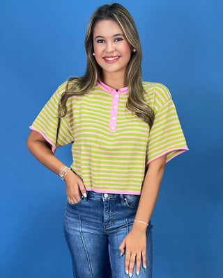 A woman modeling a short-sleeve striped henley top with lime green and white horizontal stripes and pink trim, paired with blue jeans, against a solid blue studio background.