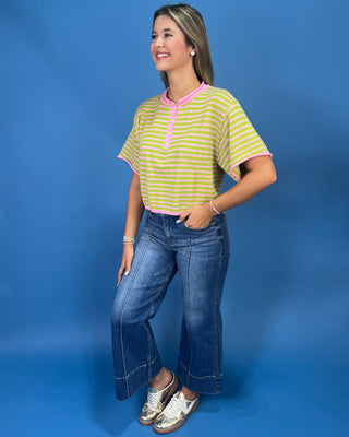 A woman modeling a short-sleeve striped henley top with lime green and white horizontal stripes and pink trim, paired with blue jeans, against a solid blue studio background.