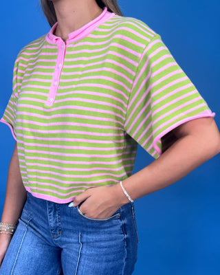 A cropped short-sleeve henley-style top with horizontal lime green and pink stripes, worn by a model with blue jeans against a solid blue background.