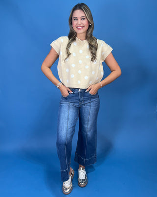 A young woman posing in a cream short-sleeve top with white polka dots, blue jeans, and silver sneakers against a solid blue studio backdrop.