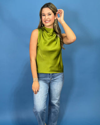 A woman modeling a sleeveless olive green satin top with a cowl neck, paired with light-wash blue jeans, standing against a solid blue studio background.