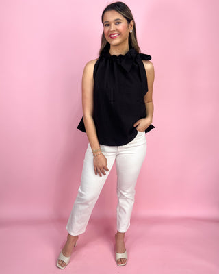 Woman wearing a black sleeveless top with a bow and white pants against a pink background