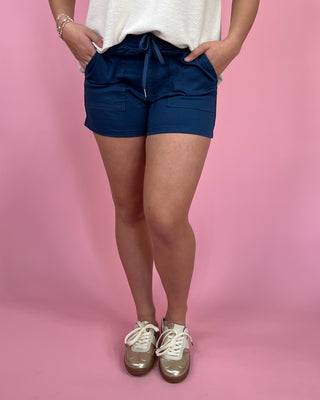 Woman wearing navy drawstring utility shorts with front pockets, styled with an ivory sleeveless top and metallic gold sneakers against a pink backdrop.