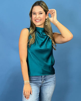 A woman modeling a sleeveless high-neck satin top in a rich teal green color, paired with light-wash blue jeans, set against a solid blue studio background.