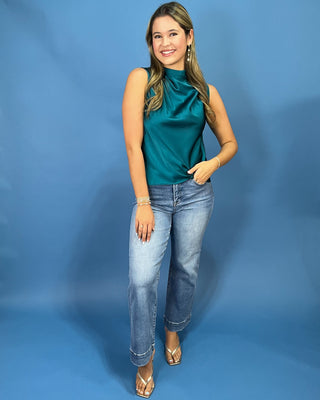 A woman modeling a sleeveless high-neck satin top in a rich teal green color, paired with light-wash blue jeans and strappy heels, standing against a solid blue studio backdrop.