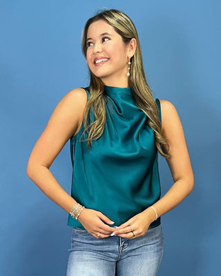 A woman with blonde-highlighted hair wearing a teal satin sleeveless top and light-wash blue jeans, standing against a solid blue studio background.