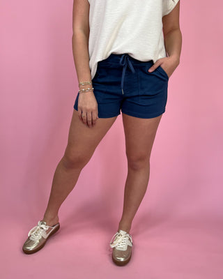 Woman wearing navy drawstring utility shorts with front pockets, styled with an ivory sleeveless top and metallic gold sneakers against a pink backdrop.