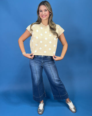 A young woman posing in a light yellow short-sleeve top with white polka dots, blue wide-leg jeans, and white sneakers against a solid blue studio background.