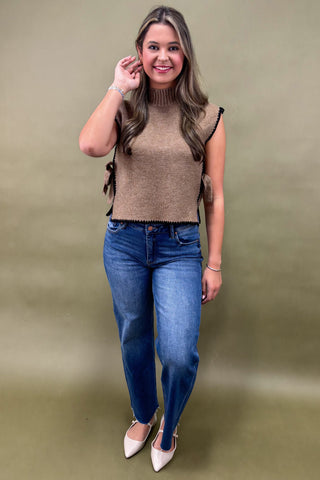 Woman wearing a brown sleeveless sweater and blue jeans against a green background