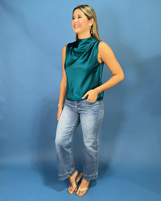 A woman modeling a sleeveless satin top in a rich teal green color, paired with light-wash blue jeans and gold sandals, standing against a solid blue studio backdrop.