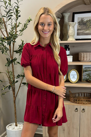 Woman in a red dress standing indoors next to a plant and shelves.