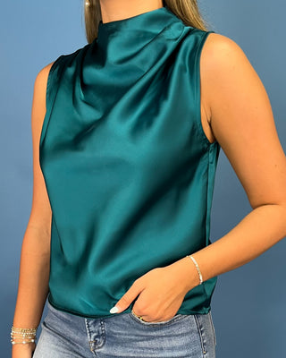 A woman wearing a sleeveless high-neck satin top in a rich teal green color, paired with blue jeans, against a solid blue background.