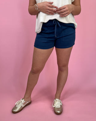 Woman wearing navy drawstring utility shorts with front pockets, styled with an ivory sleeveless top and metallic gold sneakers against a pink backdrop.