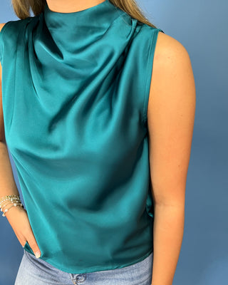 A close-up of a woman wearing a sleeveless, high-neck satin blouse in a rich teal green color, paired with light-wash blue jeans against a solid blue background.