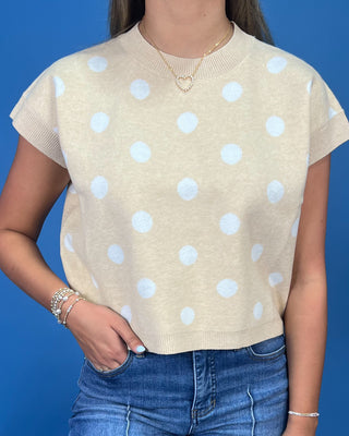 A woman wearing a beige short-sleeve knit top with white polka dots, paired with blue jeans, against a solid blue background.