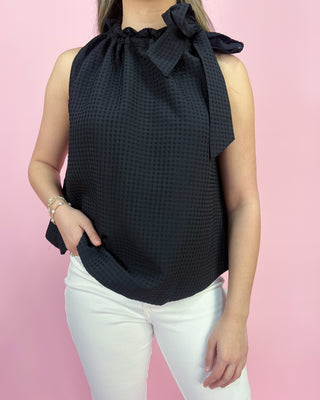 Woman wearing a black sleeveless top with a bow detail against a pink background
