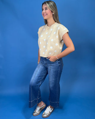 A woman modeling a beige short-sleeve knit top with white polka dots, paired with blue jeans and metallic shoes, set against a solid blue studio background.