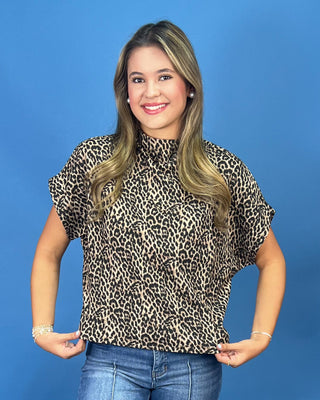 A woman with blonde-highlighted hair wearing a short-sleeve leopard-print blouse and blue jeans, standing against a solid blue studio background.