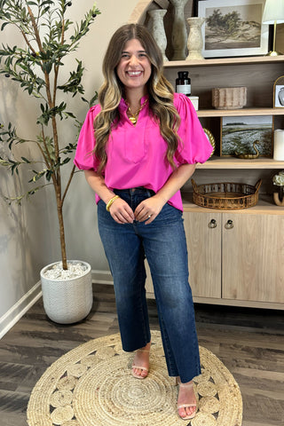 Woman in a bright pink blouse and blue jeans standing in a room with a plant and shelves.