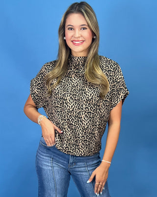 A woman modeling a short-sleeve leopard print mock neck top with blue jeans, standing against a solid blue background.