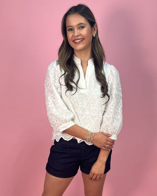 Woman wearing a white lace blouse and black shorts against a pink background