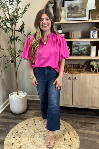 Woman in a bright pink blouse and blue jeans standing in a room with a plant and wooden cabinet.