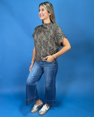 A woman modeling a short-sleeve leopard-print mock neck top with blue jeans and metallic sneakers, standing against a solid blue studio backdrop.