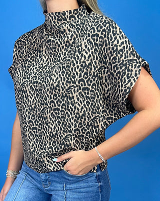 A woman wearing a leopard print short-sleeve blouse with a high mock neck, paired with blue jeans, set against a solid blue background.