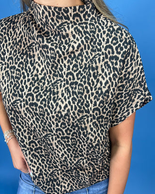 A person wearing a short-sleeve leopard print blouse with a high mock neck, paired with blue jeans, set against a solid blue background.