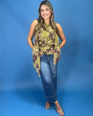 A woman modeling a sleeveless olive green and brown floral print top with blue jeans, standing against a solid blue studio backdrop.