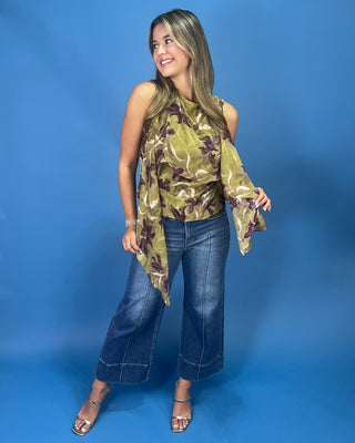 A woman modeling a sleeveless olive green and brown floral print top with wide-leg blue jeans and silver sandals, standing against a solid blue studio backdrop.
