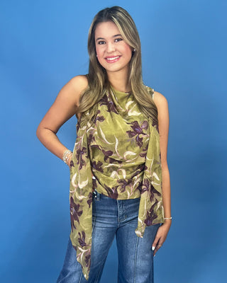 A woman modeling a sleeveless olive green top with a brown and beige floral pattern, paired with blue jeans, against a solid blue studio background.