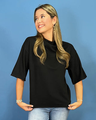 A woman with blonde-highlighted hair wearing a black short-sleeve mock neck top and light blue jeans, standing against a solid blue background.