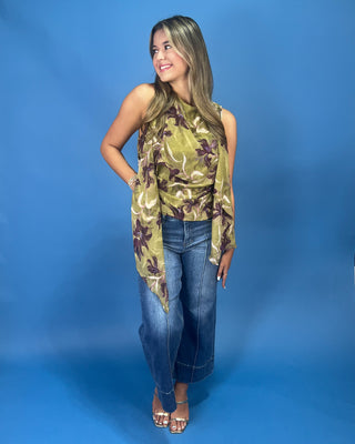 A woman modeling a sleeveless olive green top with a brown and purple floral pattern, paired with blue jeans, standing against a solid blue studio backdrop.