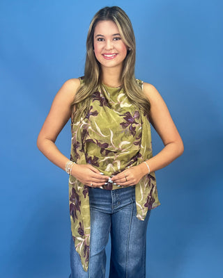 A woman modeling a sleeveless olive green top with a brown and purple floral pattern, paired with blue jeans, against a solid blue studio background.