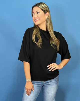 A woman modeling a black short-sleeve mock neck t-shirt with light wash blue jeans, standing against a solid blue backdrop.