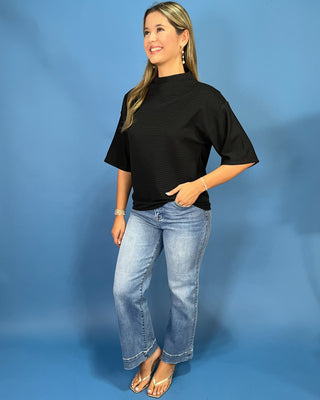 A woman modeling a black short-sleeve mock neck t-shirt with wide sleeves, paired with light-wash blue jeans and gold sandals, standing against a solid blue studio backdrop.