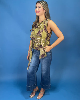 A woman modeling a sleeveless olive green and brown floral print top with a cowl neck and tie detail, paired with blue jeans and metallic sandals, against a solid blue studio background.