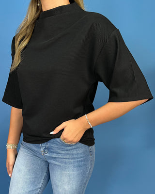 A woman wearing a black short-sleeve ribbed top with a mock neck, paired with light blue jeans, standing against a solid blue background.
