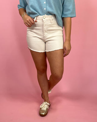 Woman wearing white high-waisted denim shorts with a raw hem, styled with a light blue button-front top and metallic gold sneakers against a pink backdrop.