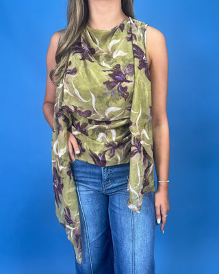 A woman wearing a sleeveless olive green draped top with a purple and white floral print, paired with blue jeans, standing against a solid blue background.