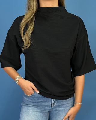 A woman wearing a black short-sleeve ribbed mock neck top and light blue jeans, standing against a solid blue background.