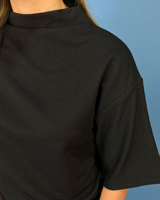 Close-up of a person wearing a black ribbed short-sleeve top against a solid blue background.