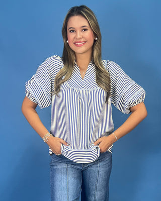 A woman modeling a short-sleeve striped blouse with puff sleeves and a V-neckline, paired with blue jeans, standing against a solid blue background.